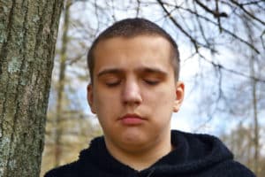 Close up of a young man in a dark sweatshirt standing in a wooded area with his eyes closed.