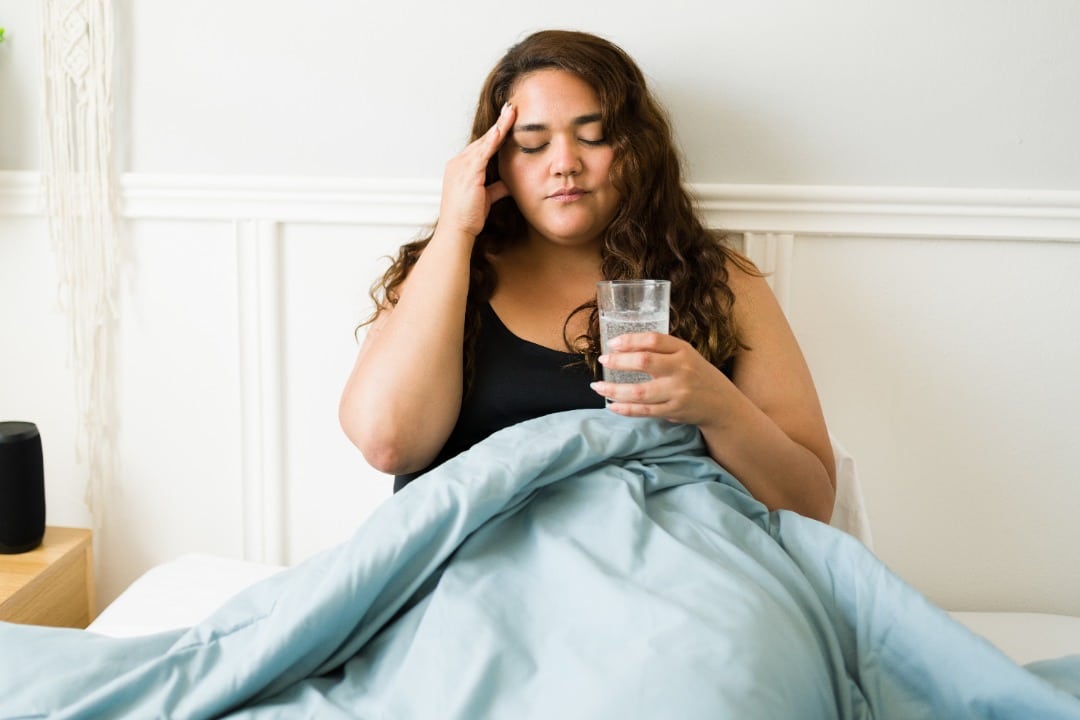 Woman sitting up in bed with a glass of water. She's holding her head because of a hangover headache.