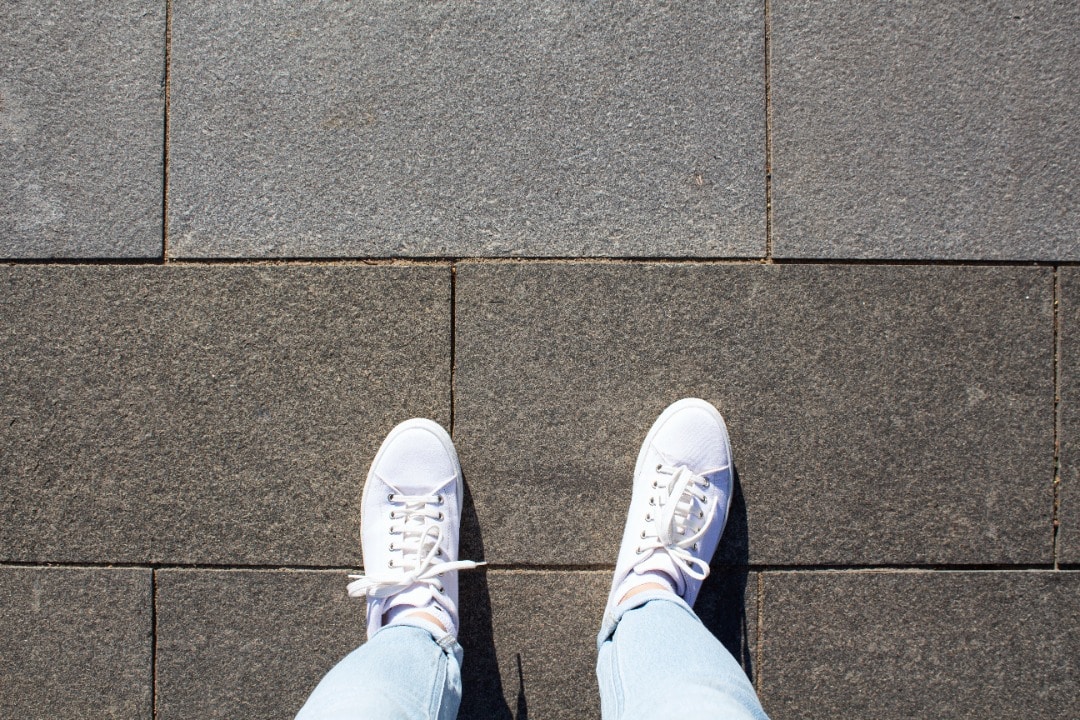 Point of view is looking down at feet in white tennis shoes standing on cement pavers.