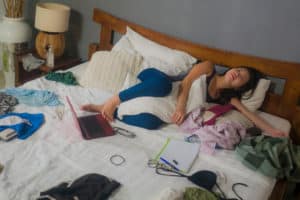 Young woman sits on her bed surrounded by a mess.